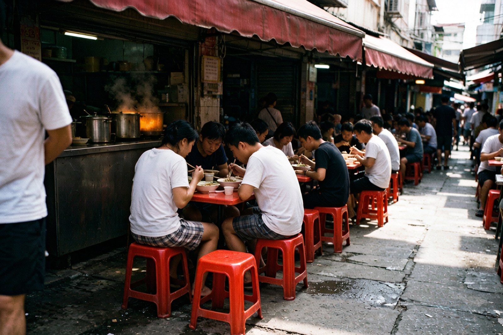 A street corner at the entrance to the market, plastic stools out, customers eating noodle bowls under a red awning