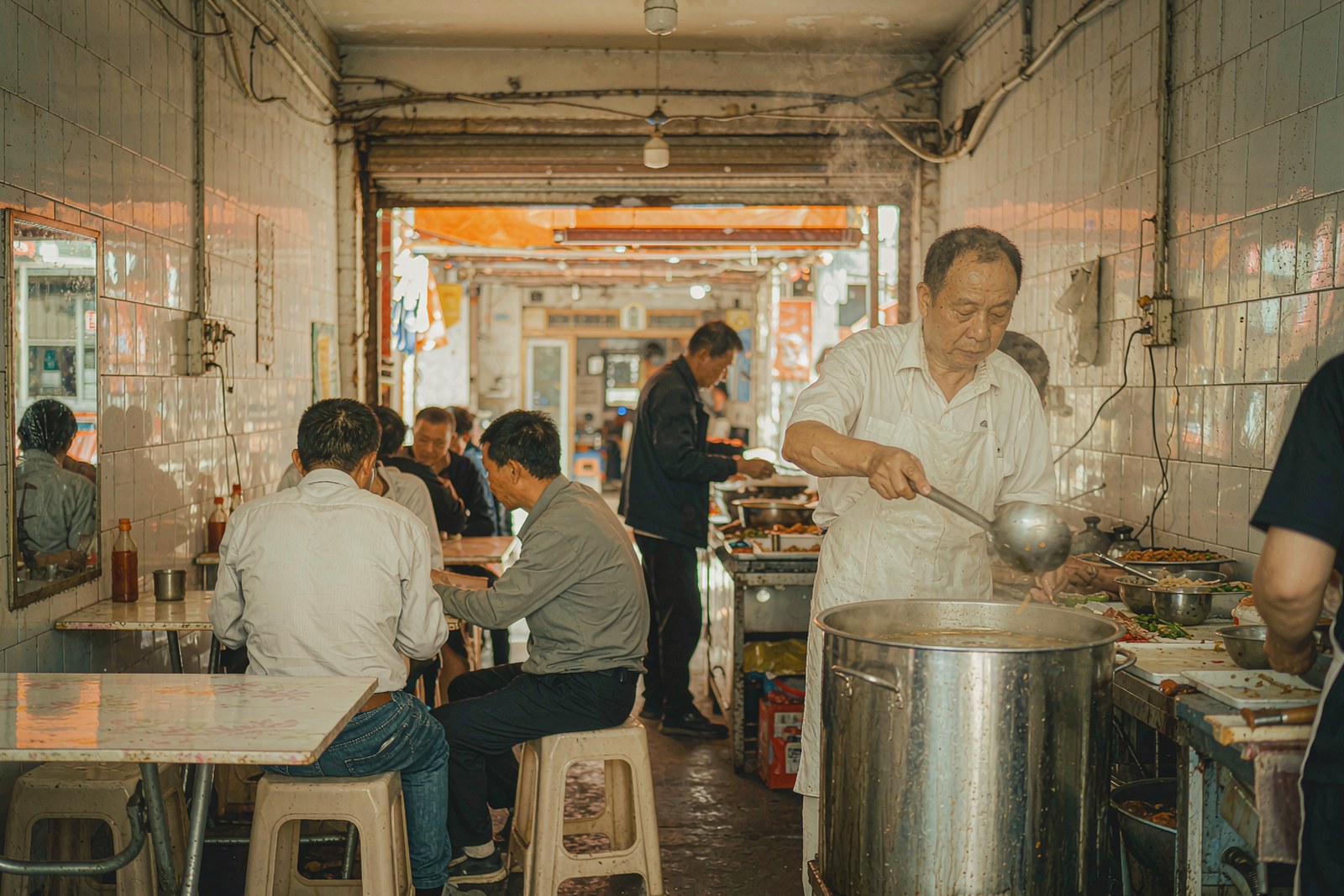 A cook ladles bone broth into bowls at the back counter as customers eat at low tables