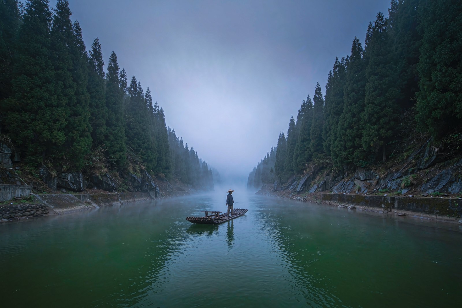 Low mist lying on a green river between forested banks, a bamboo raft drifting through