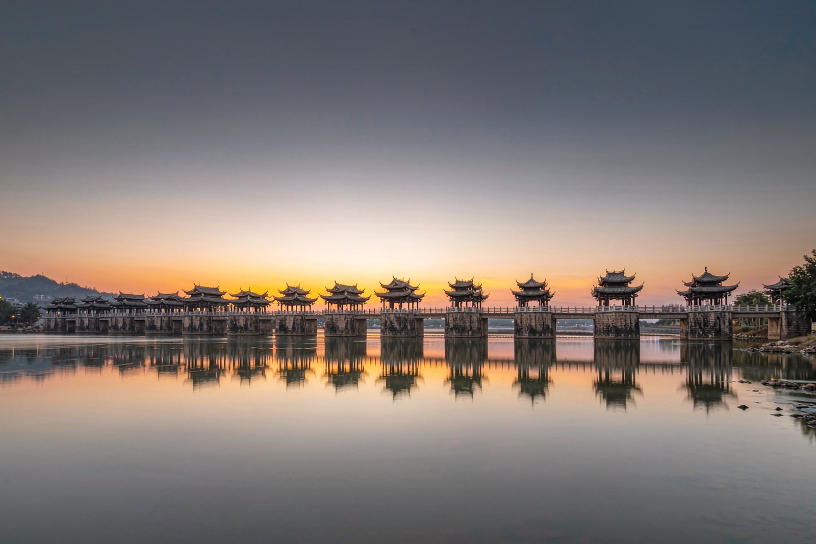A long stone-pier bridge with pavilions crossing a wide river at dusk