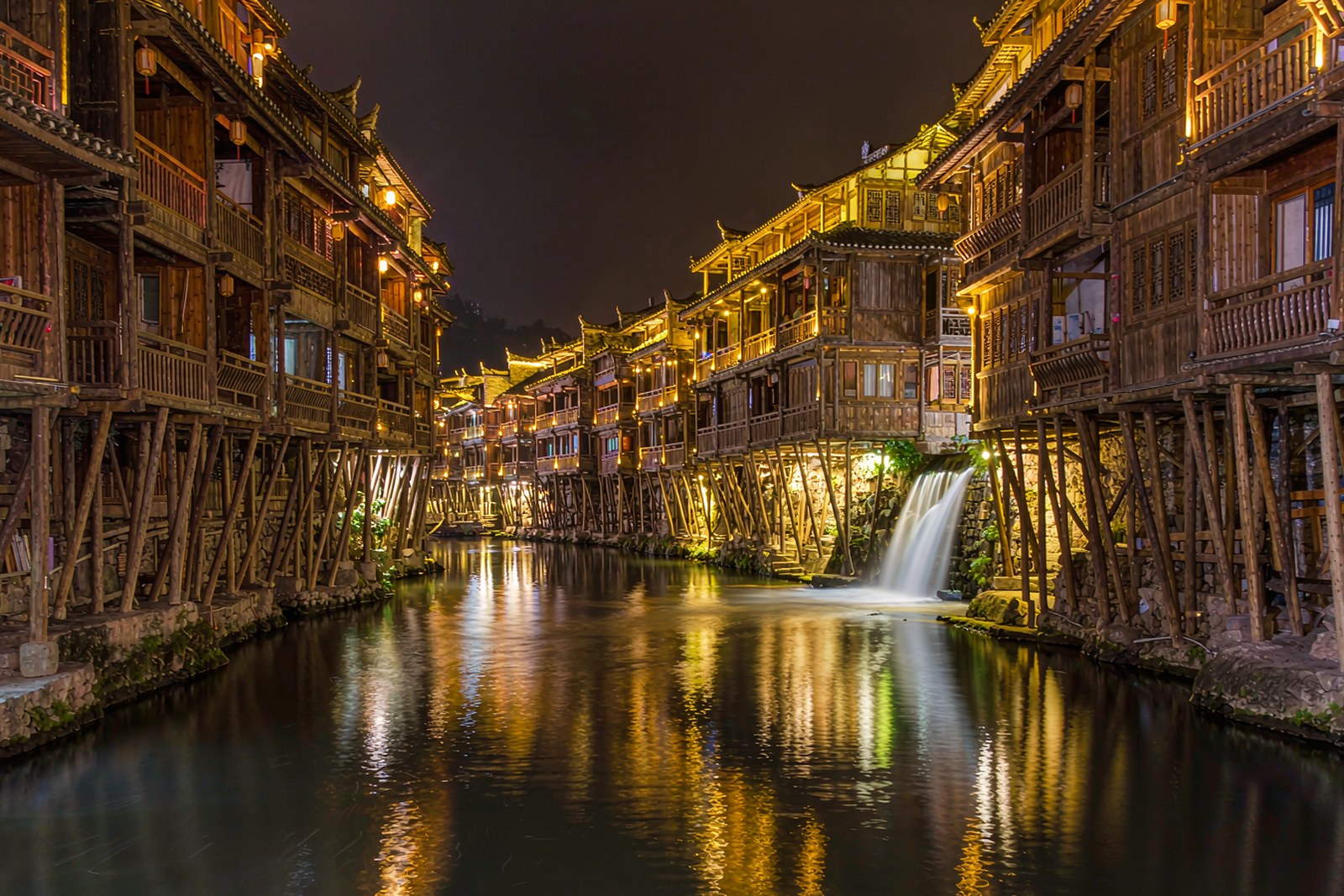 Timber Tujia houses on stilts at night, lit above a waterfall